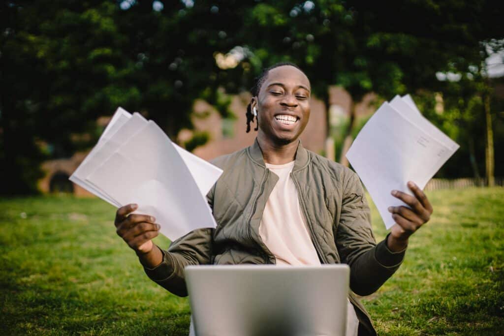 A man celebrating his first freelance writing assignment. He is sitting at a laptop and holding paper in both hands.